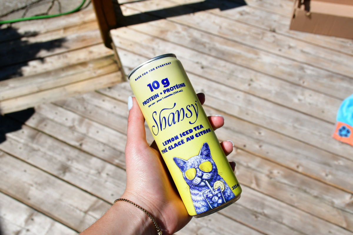 A hand holding a can of Shansy lemon iced tea protein drink against a wooden deck background.